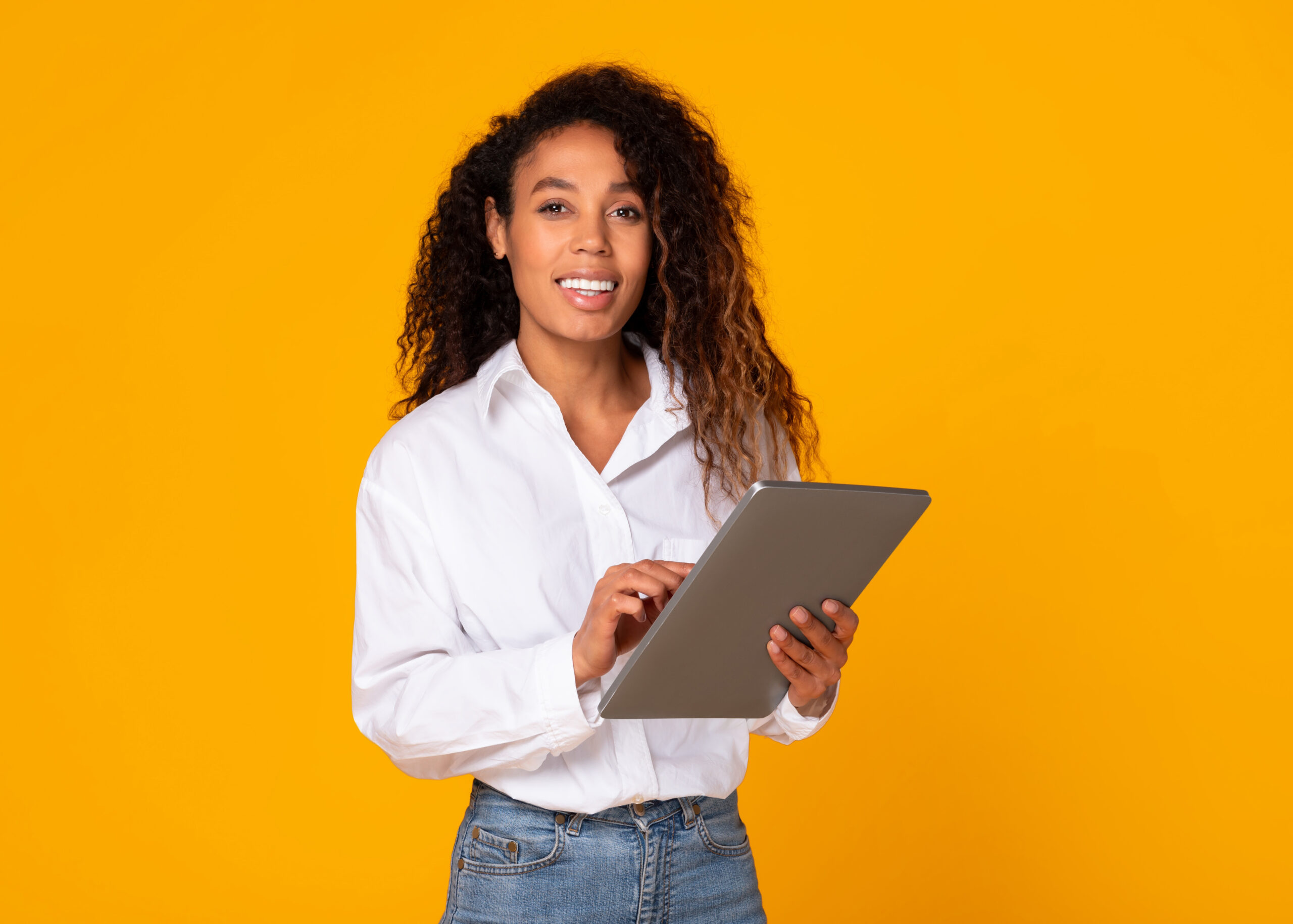 Cheerful African American lady engaging with digital tablet for freelance work in studio on yellow background. Lady in casual white shirt happily websurfing while posing with portable PC